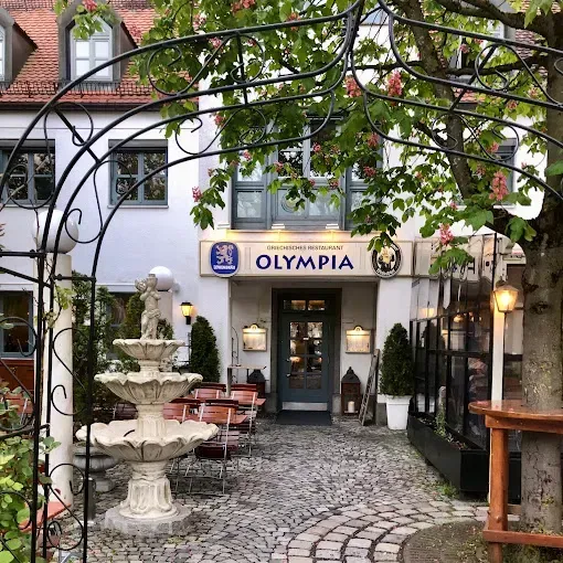 Eingang zum Restaurant „Olympia“ mit Brunnen, Tischen und einem Baumbogen.