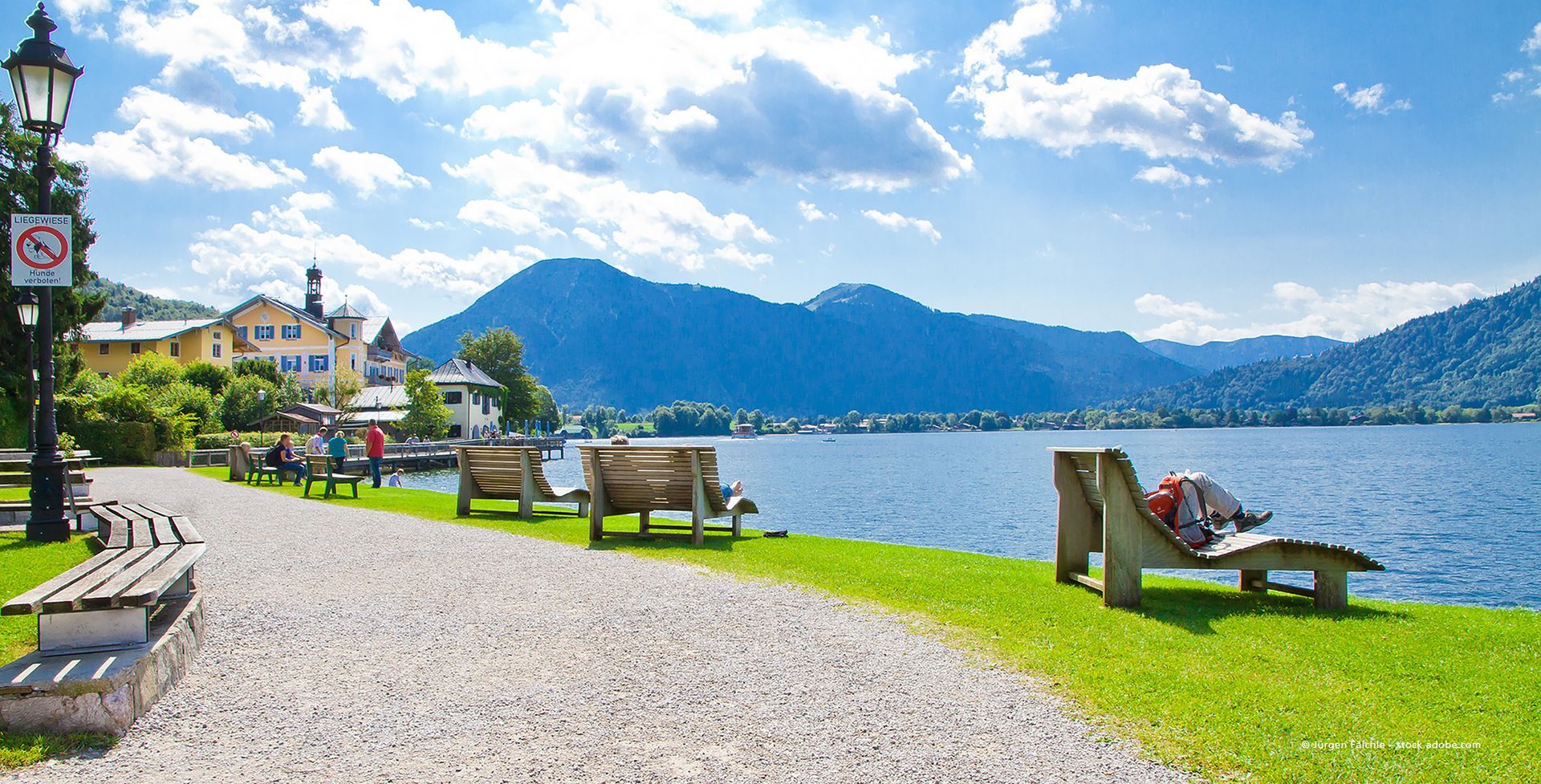 Malerischer Uferweg mit Bänken, Berge in der Ferne, blauer Himmel und Wasser.