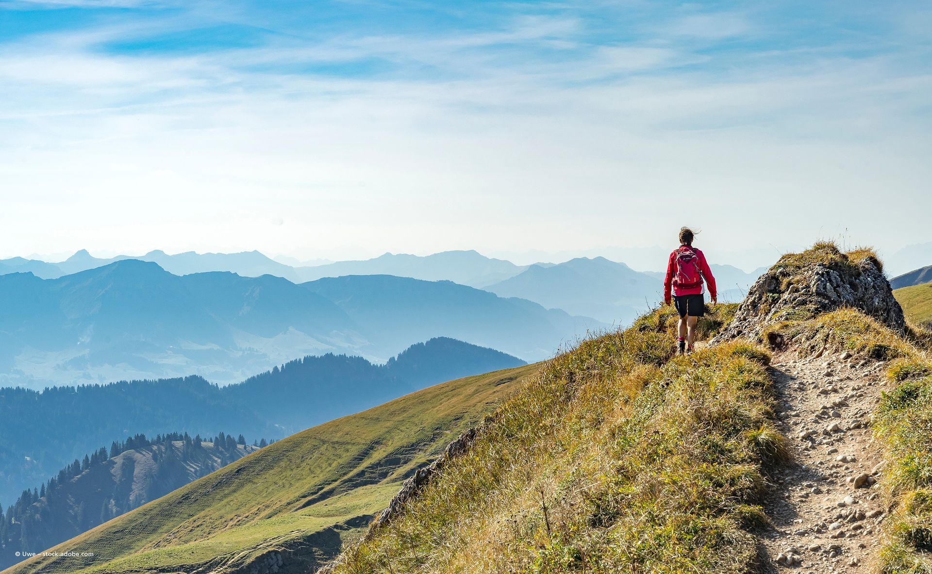 Eine Person wandert auf einem grasbewachsenen Bergkamm entlang, im Hintergrund erheben sich blaue