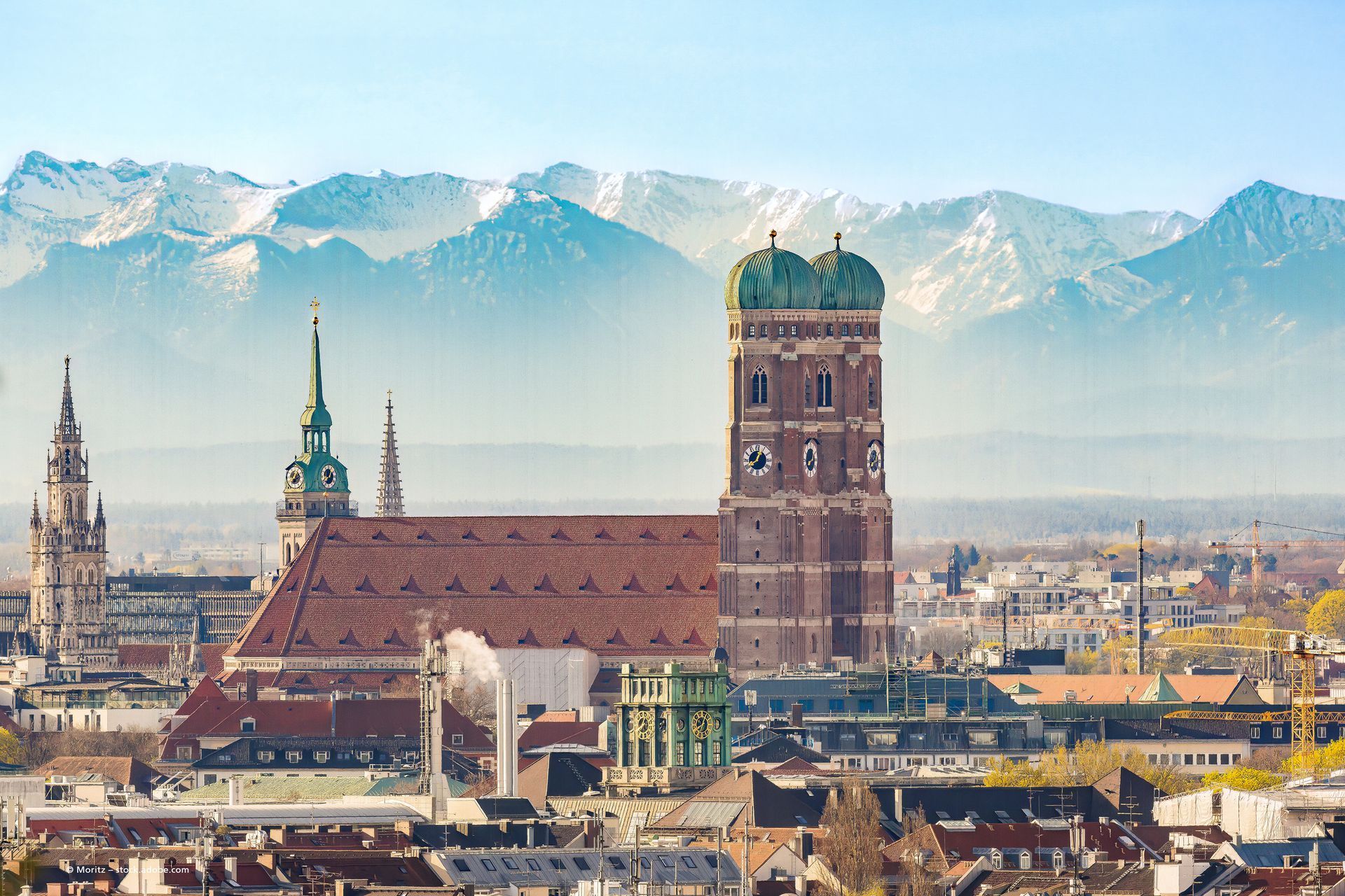 Die Münchner Skyline mit der Frauenkirche und den schneebedeckten Alpen im Hintergrund.