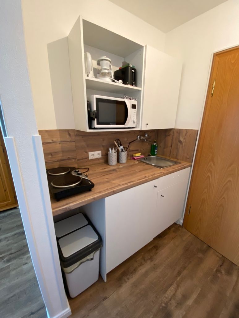 Kleine Küche mit weißen Schränken, Mikrowelle, Spüle und Mülleimer, mit Arbeitsplatte und Boden in Holzoptik.