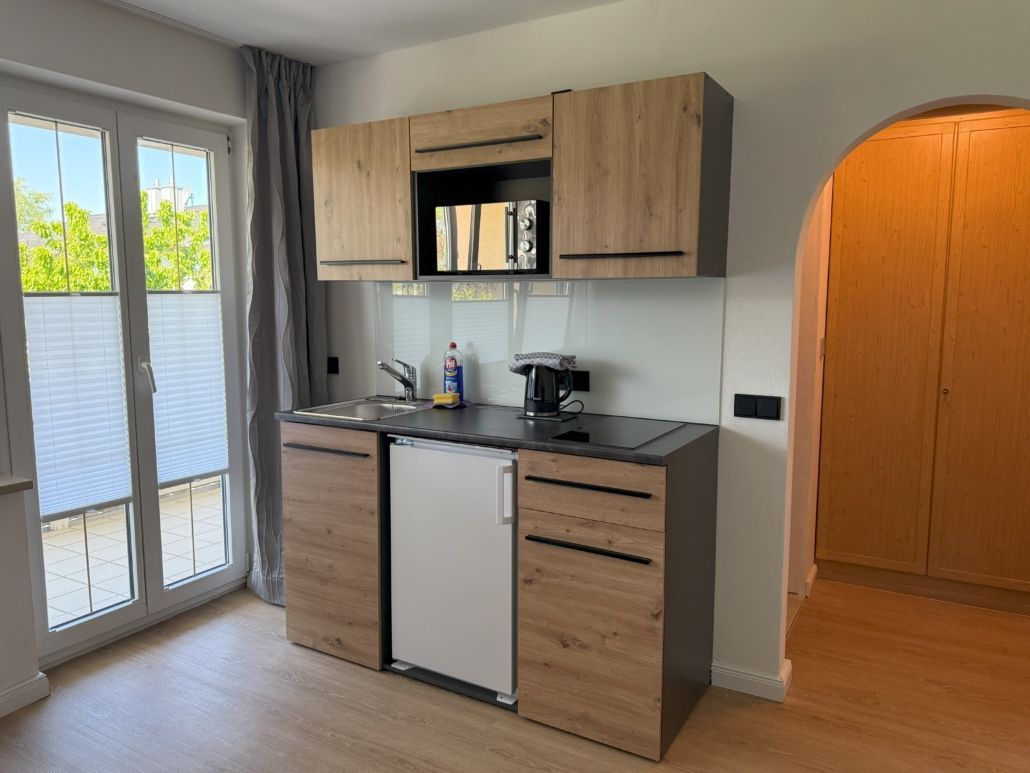 Kleine Küchenzeile mit Holzschränken, Spüle, Minikühlschrank und Mikrowelle. Eine Tür führt in einen weiteren Raum.