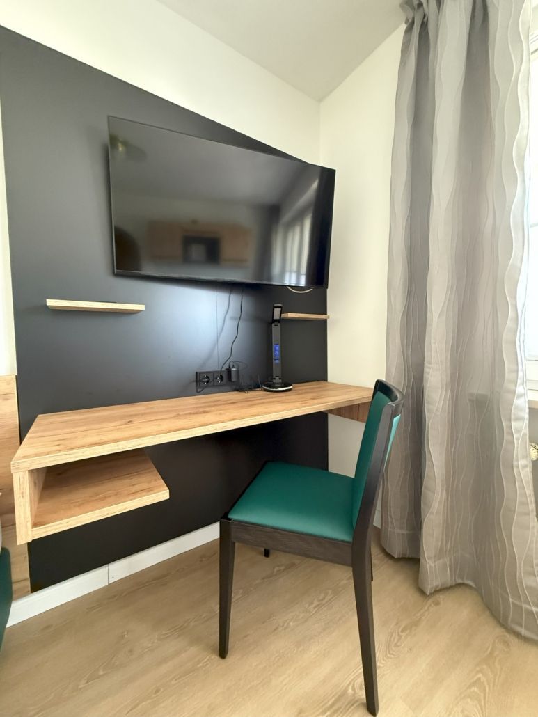 Schreibtisch mit an der Wand montiertem Fernseher, kleinen Regalen und einem türkisfarbenen Stuhl vor einer schwarzen Wand.