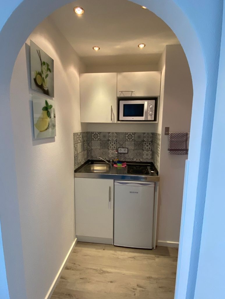 Kleine Küchenzeile mit weißen Schränken, Mikrowelle und Minikühlschrank hinter einem Bogeneingang.