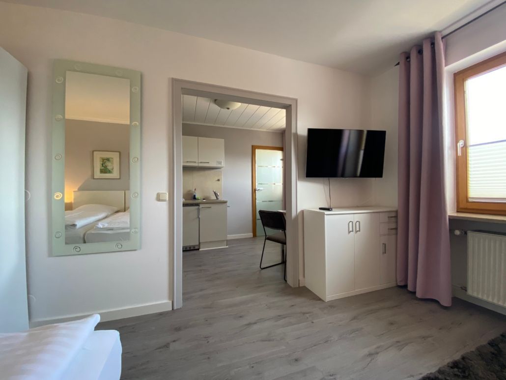 Innenansicht eines Studio-Apartments mit grauem Boden, Küche und Bett. Über einem weißen Schrank hängt ein Fernseher.