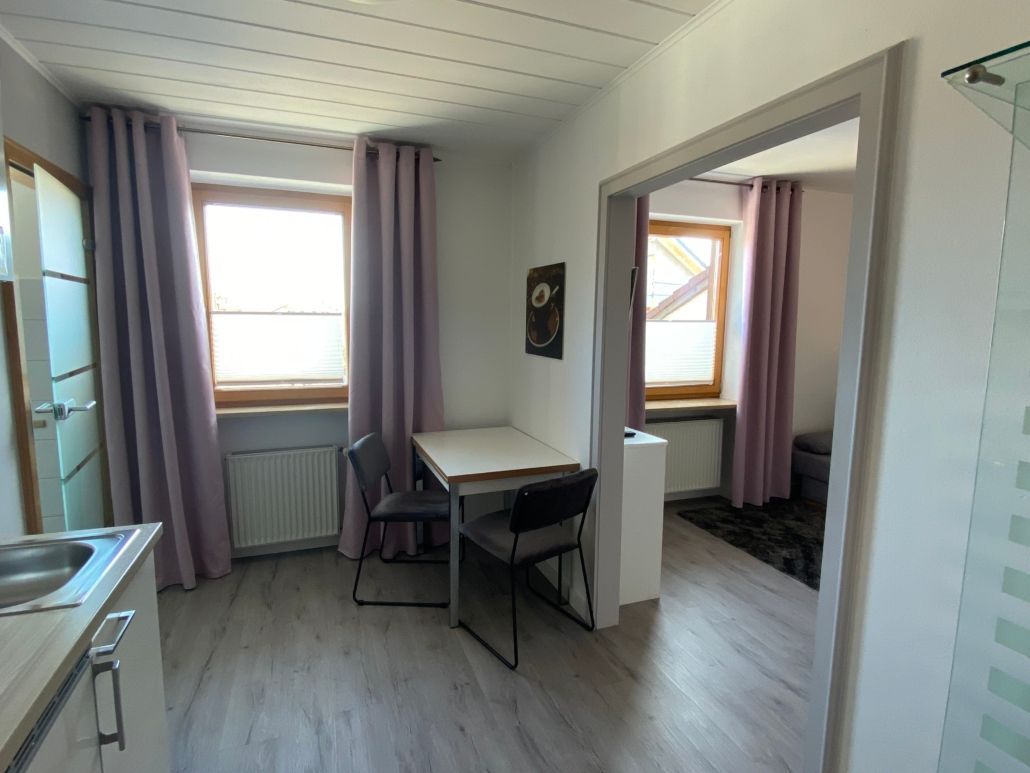 Kleiner Raum mit Tisch, zwei Stühlen, zwei Fenstern mit violetten Vorhängen und einer Tür zu einem anderen Raum.