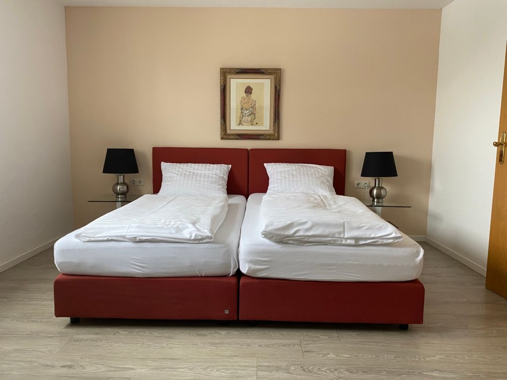 Rotes Bett mit zwei separaten Matratzen, zwei schwarze Lampen und ein gerahmtes Bild an der beigen Wand.