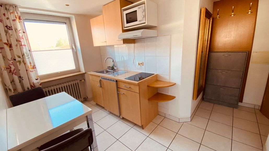 Kleine Küchenzeile in einem Studio-Apartment mit hellen Holzschränken, Mikrowelle und kleinem Tisch am Fenster.