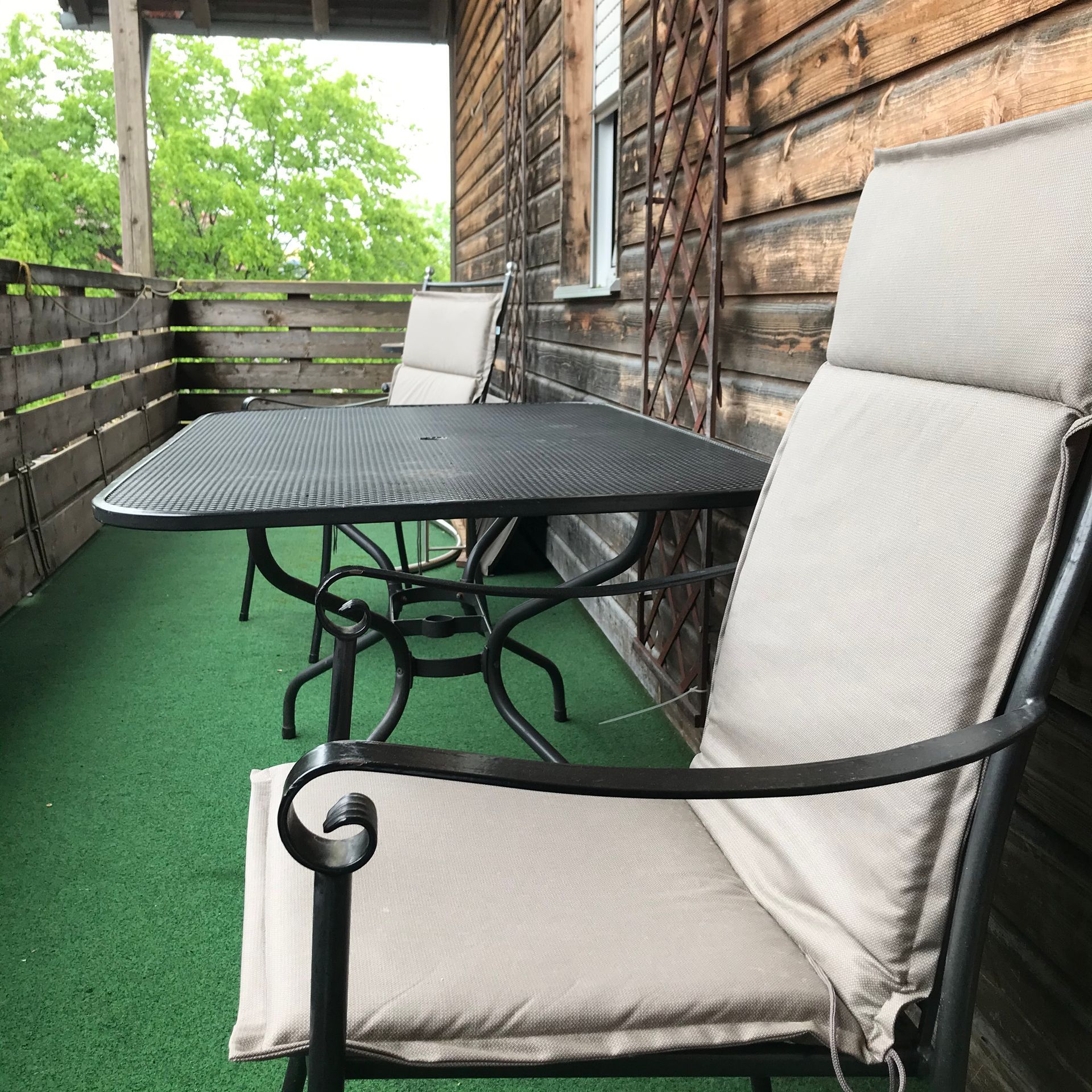 Balkon mit Metalltisch und -stühlen, Holzverkleidung, grünem Rasen und Bäumen im Hintergrund.