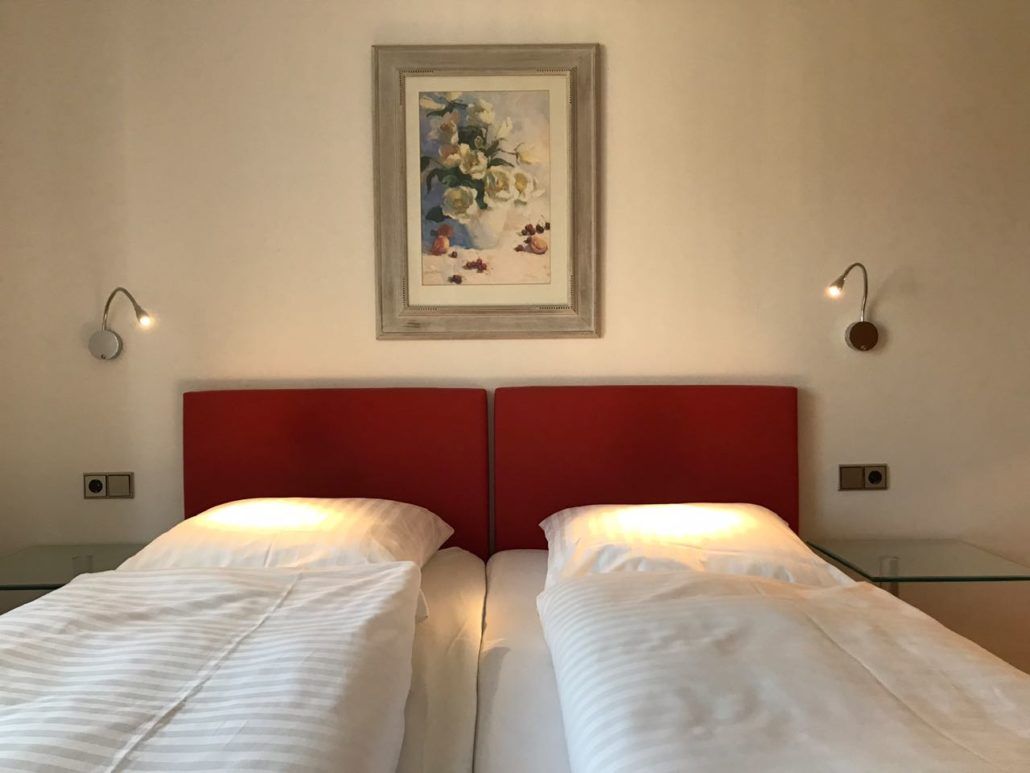 Zwei Einzelbetten mit weißer Bettwäsche, roten Kopfteilen und Wandleuchten. Darüber hängt ein Kunstwerk.