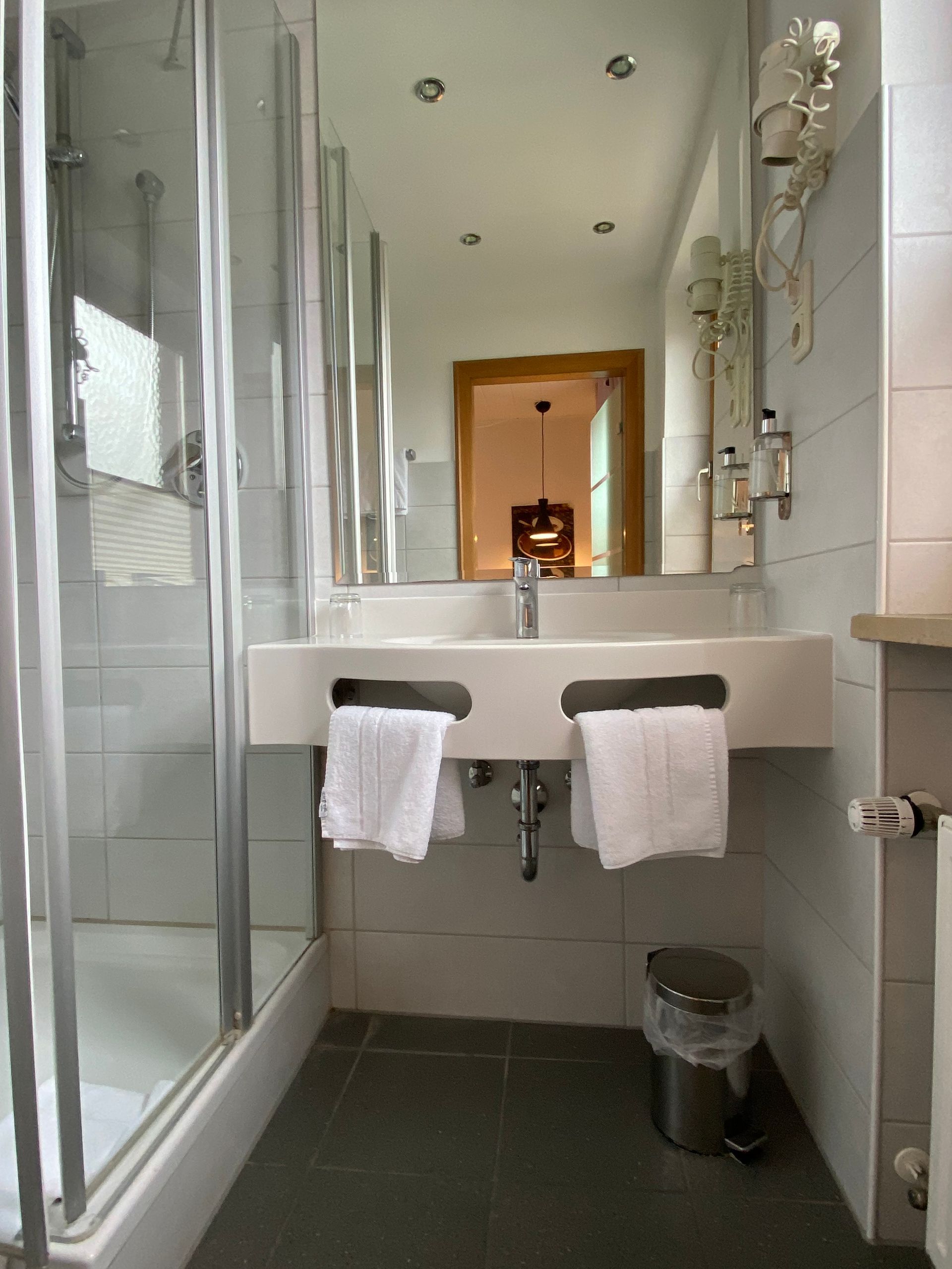 Badezimmer mit Dusche, Waschtisch mit zwei Waschbecken, Handtüchern, Spiegel und Mülleimer.