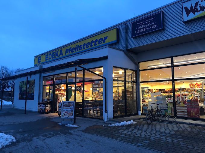 Außenansicht des Edeka-Supermarkts in der Abenddämmerung; beleuchtete Fenster, Namensschild, Fahrrad draußen geparkt.