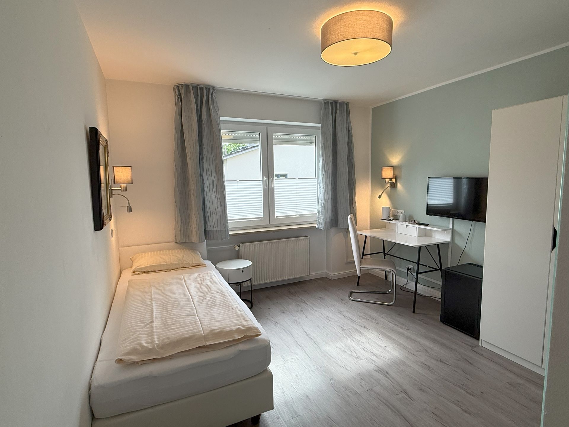 Kleines, helles Hotelzimmer mit Einzelbett, Schreibtisch, Fernseher und Fenster mit Vorhängen.