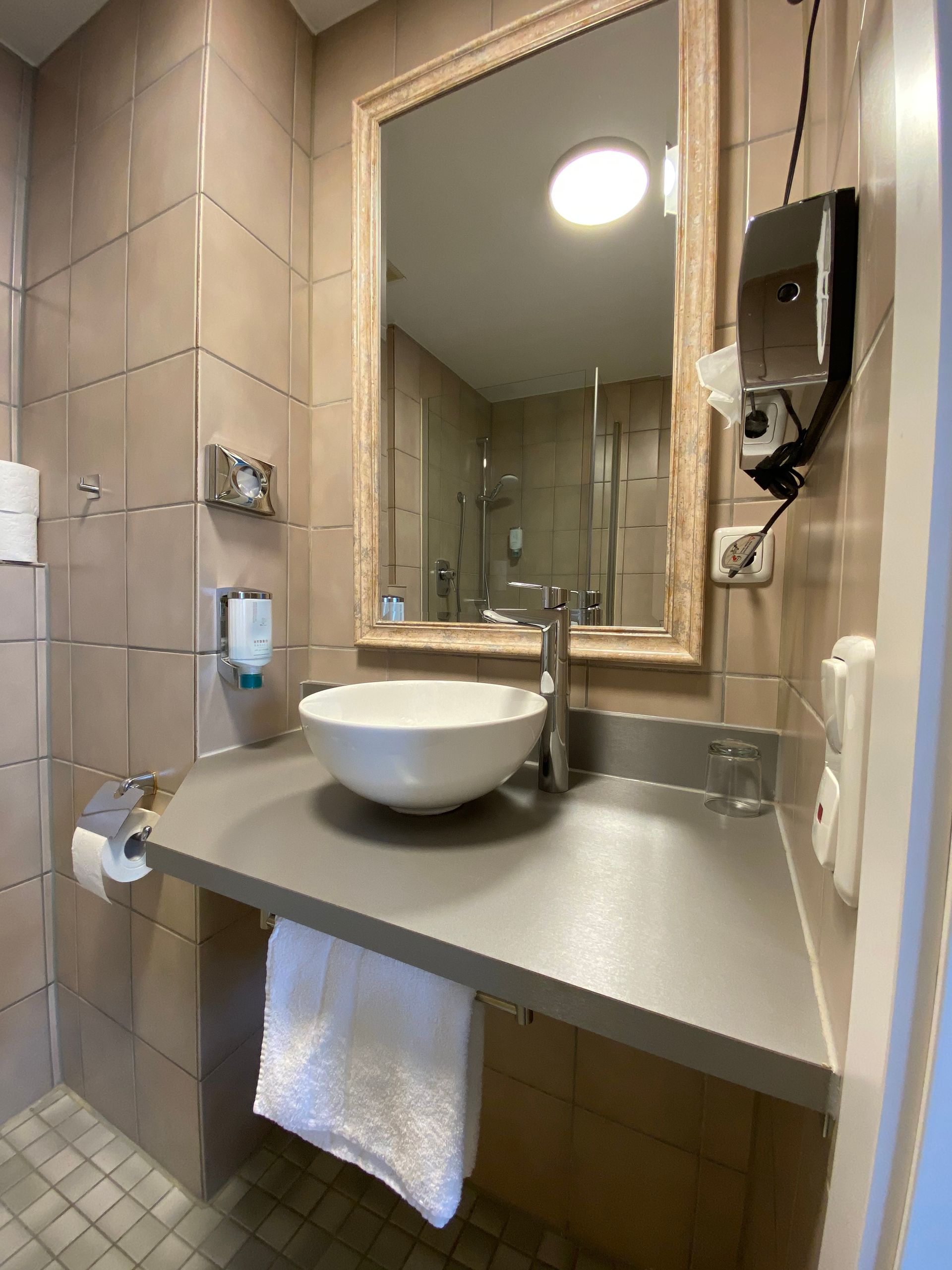 Badezimmer mit Standwaschbecken, Spiegel, Handtuchhalter und Seifenspender. Hellbraune Fliesen und eine helle Deckenleuchte.