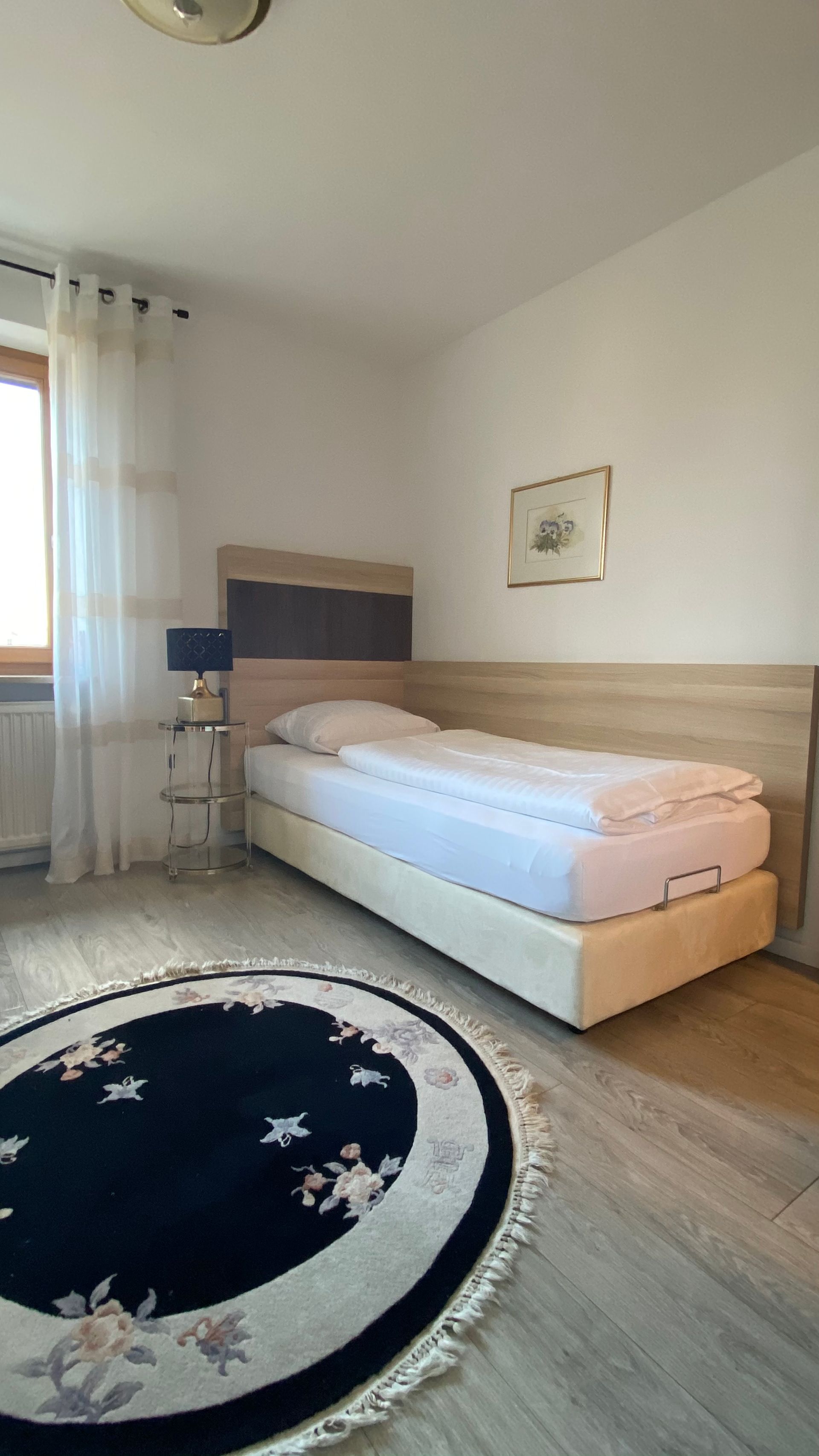 Schlafzimmer mit Einzelbett, Nachttisch mit Lampe, rundem Teppich und Fenster.