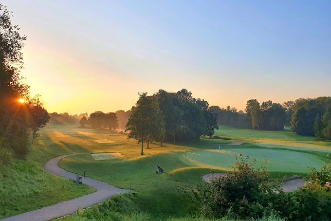 Sonnenaufgang über einem Golfplatz. Grüne Fairways, Bäume und ein Weg im Morgennebel.