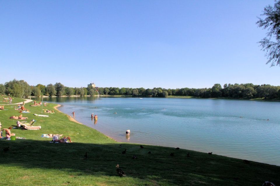 Szene am See: Menschen sonnen sich und schwimmen. Blaues Wasser, grünes Gras, Bäume und klarer Himmel.