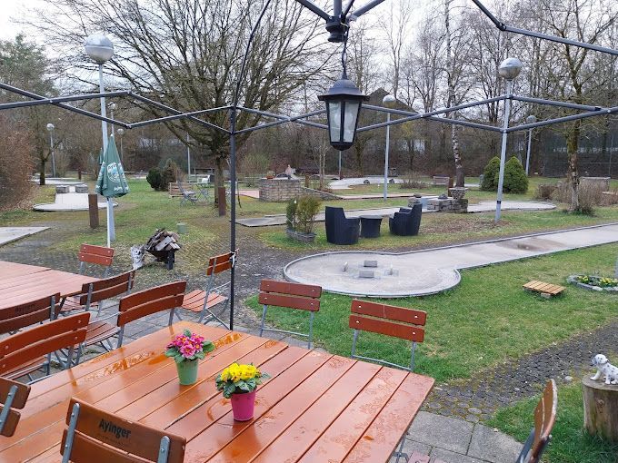 Minigolfanlage mit Tischen und Stühlen unter einer Metallpergola; ein Springbrunnen und kleine Gebäude auf dem Green.