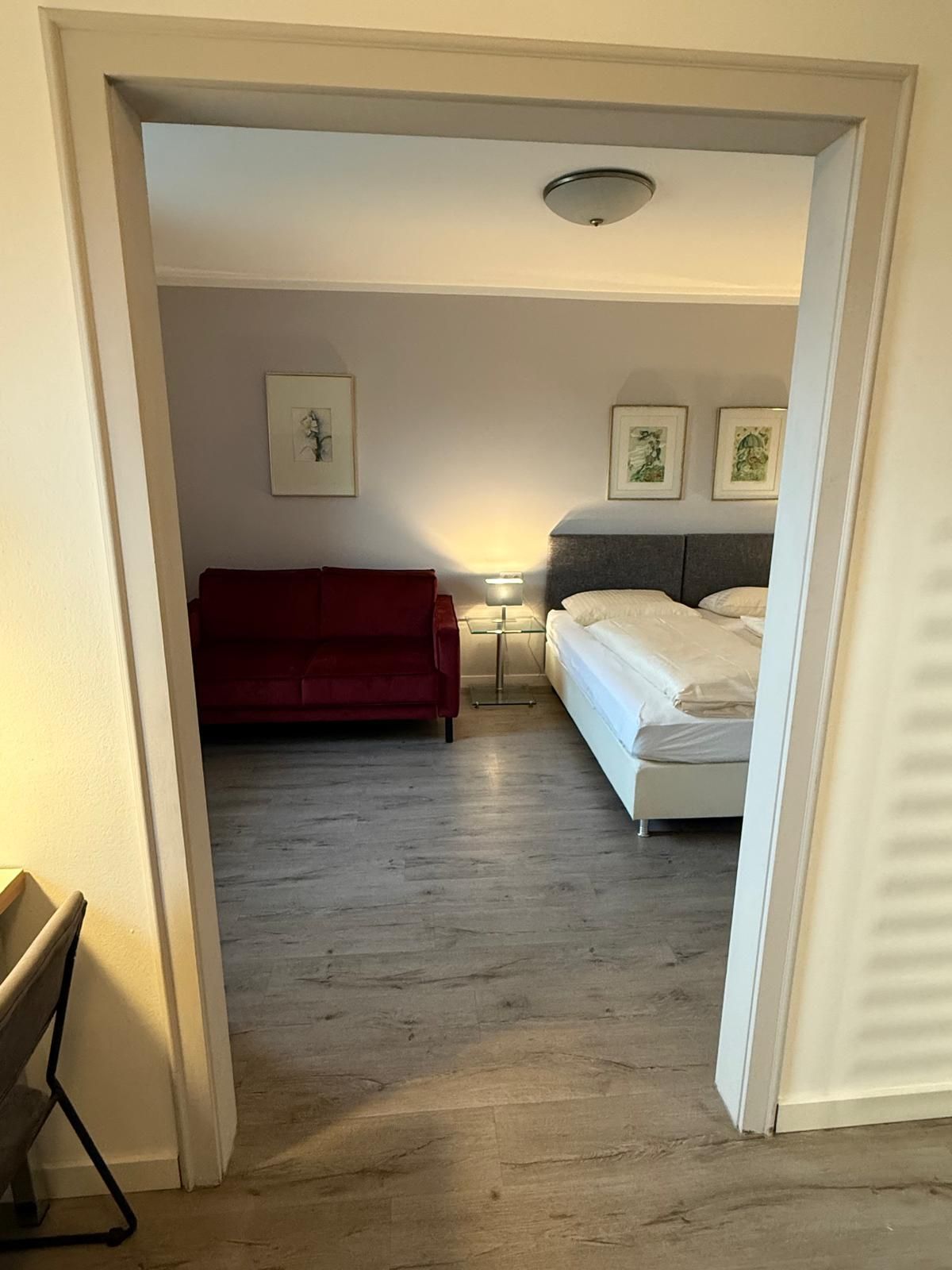 Schlafzimmer mit Bett, Sofa, Bildern und Teppich. Graue Wände und grauer Boden. Zwei Lampen sorgen für warmes Licht.