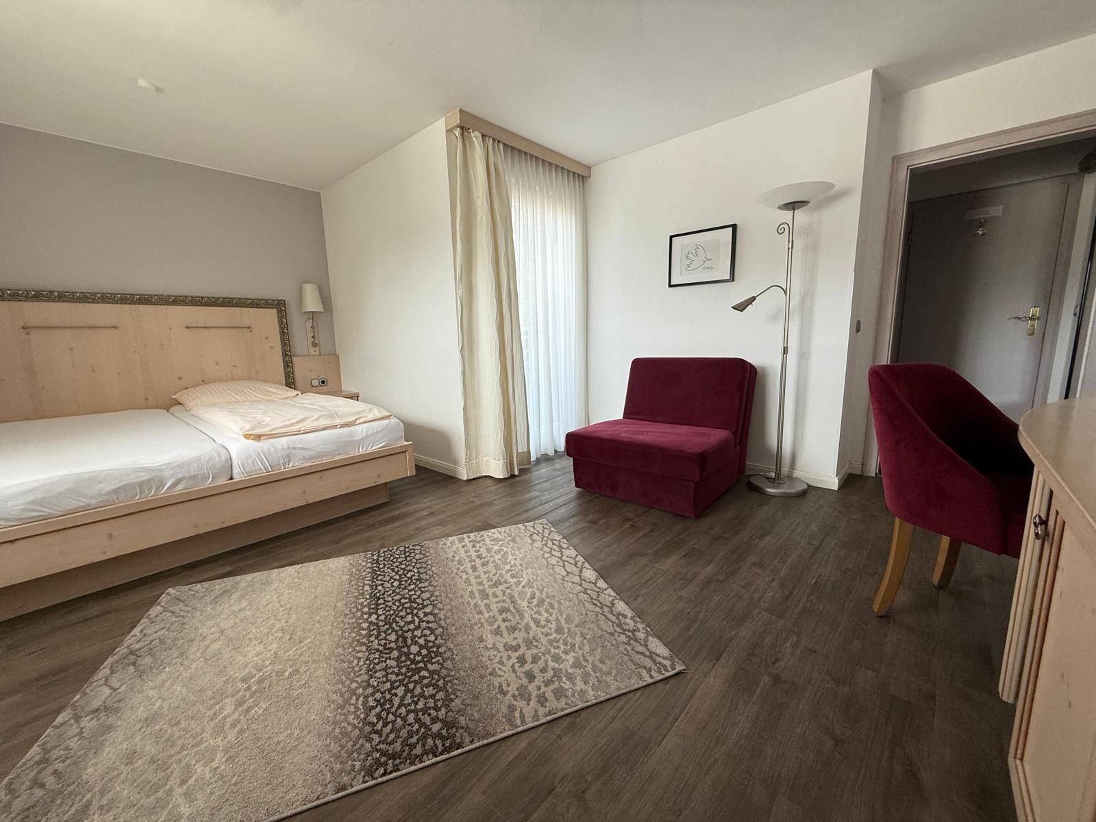 Schlafzimmer mit Bett, Stuhl, Schreibtisch, Fernseher und Kleiderschrank; neutrale Farben.