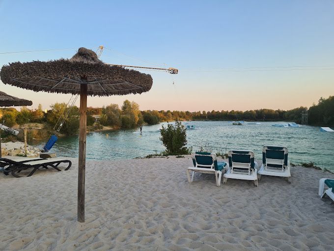 Strand mit Strohschirmen, Liegestühlen und Wassersportmöglichkeiten an einem See unter dämmerigem Abendhimmel.