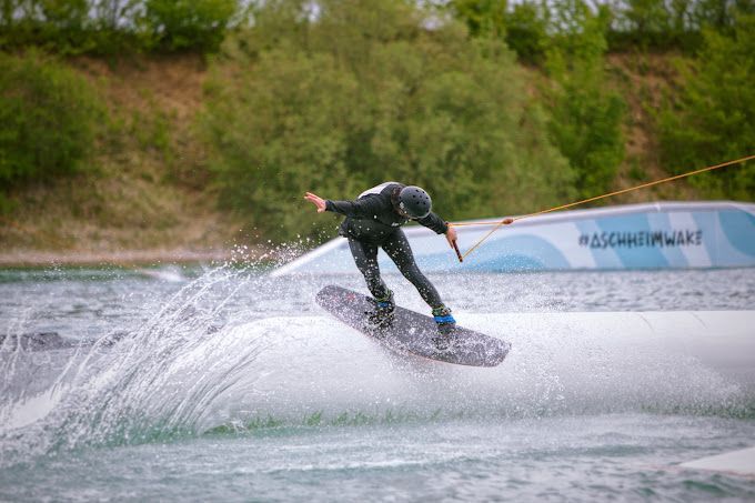 Wakeboarder im schwarzen Neoprenanzug gleitet über das Wasser und verursacht einen Spritzer.