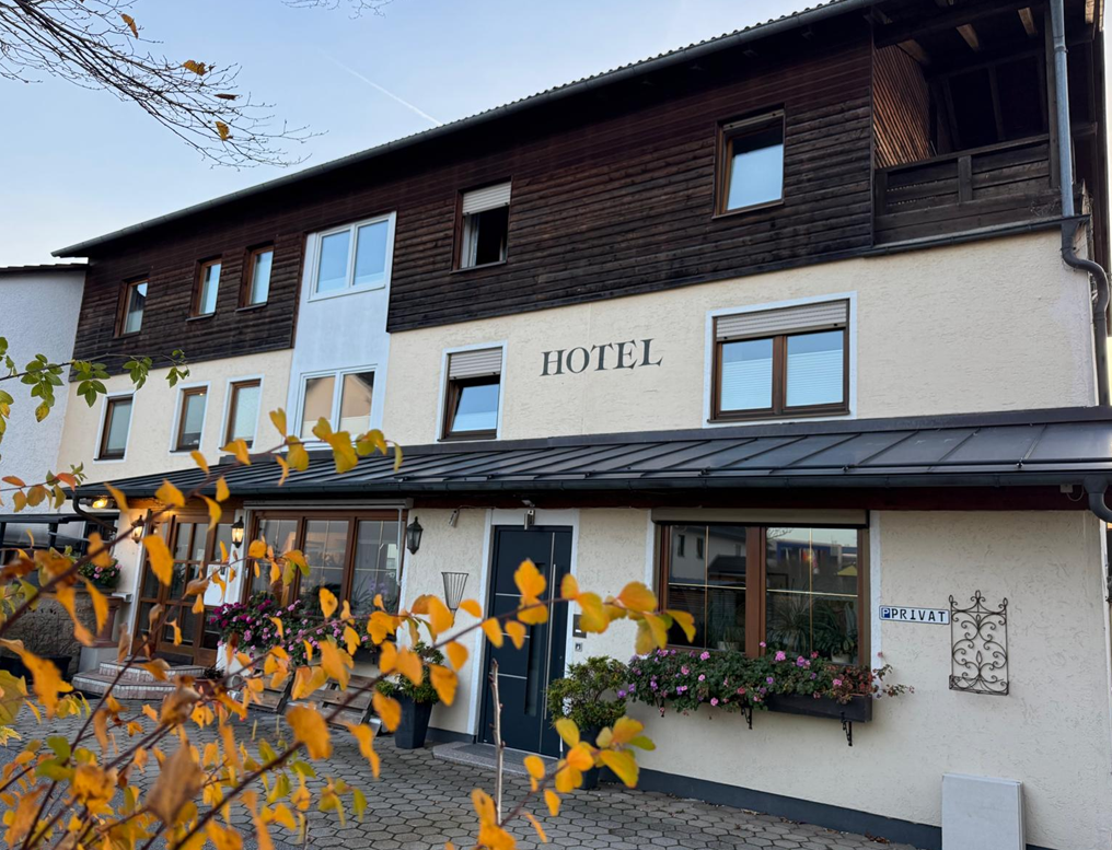 Hotelaußenansicht mit weißer Fassade, braunem Holzobergeschoss und dunklem Dach. Gelbe Blätter im Vordergrund.