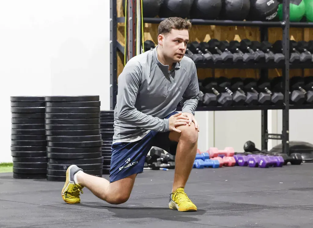 Hombre en el gimnasio haciendo una zancada con las manos sobre las rodillas. Pesas apiladas y mancuernas al fondo.