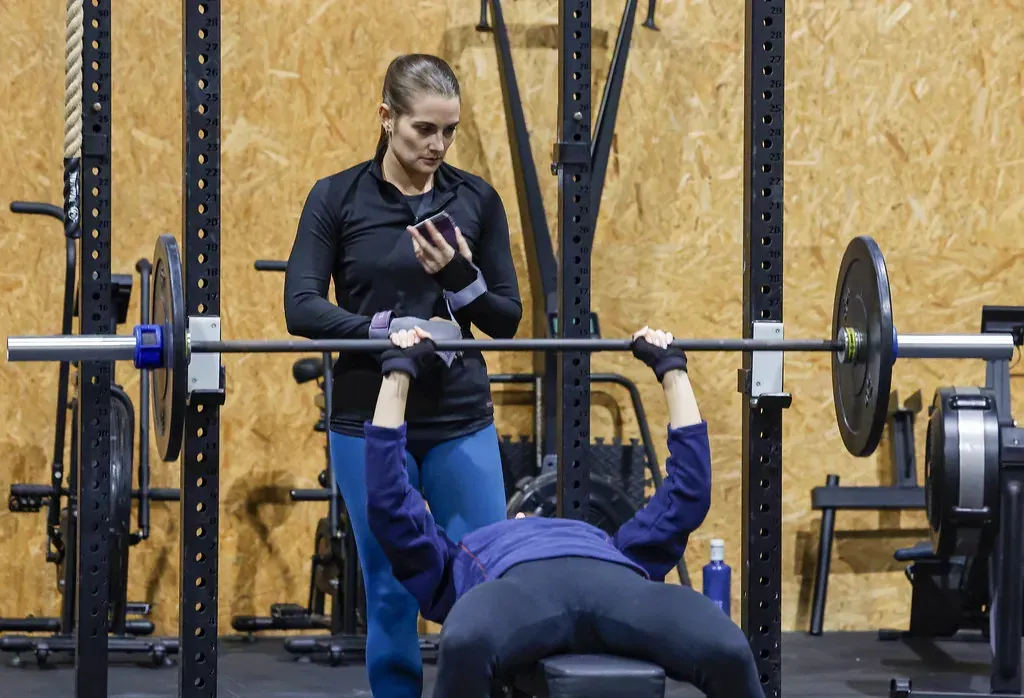 Una mujer hace press de banco con barra con un observador en un gimnasio.