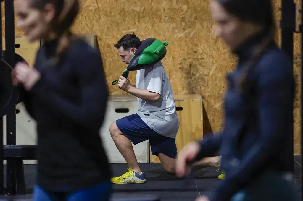 Un hombre en el gimnasio hace zancadas con una bolsa de deporte verde. Otras personas hacen ejercicio al fondo.