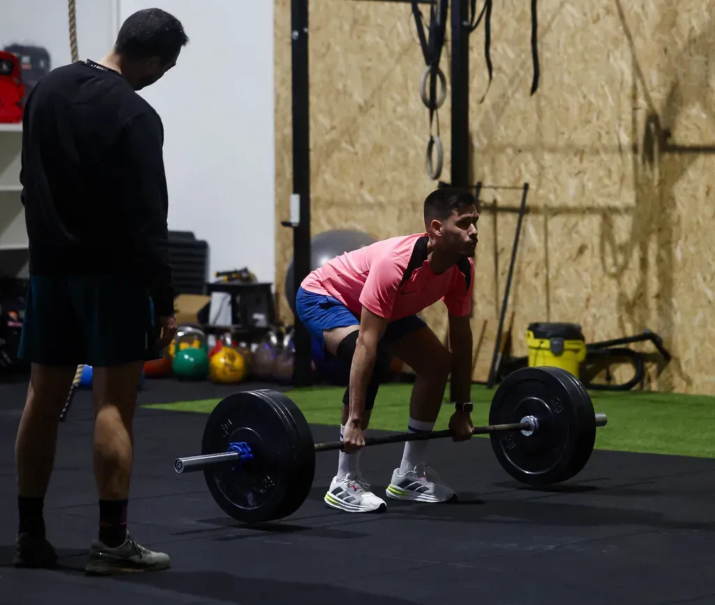 Un hombre levanta una barra con un instructor observándolo en un gimnasio.