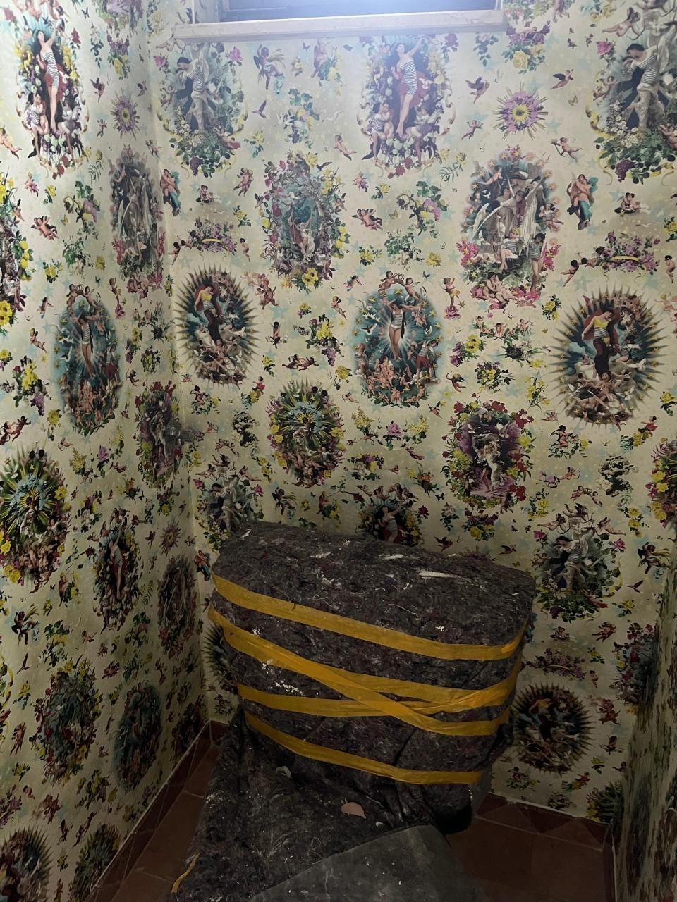 In einem Raum mit geblümter Tapete ist eine Toilette am Boden festgeklebt.