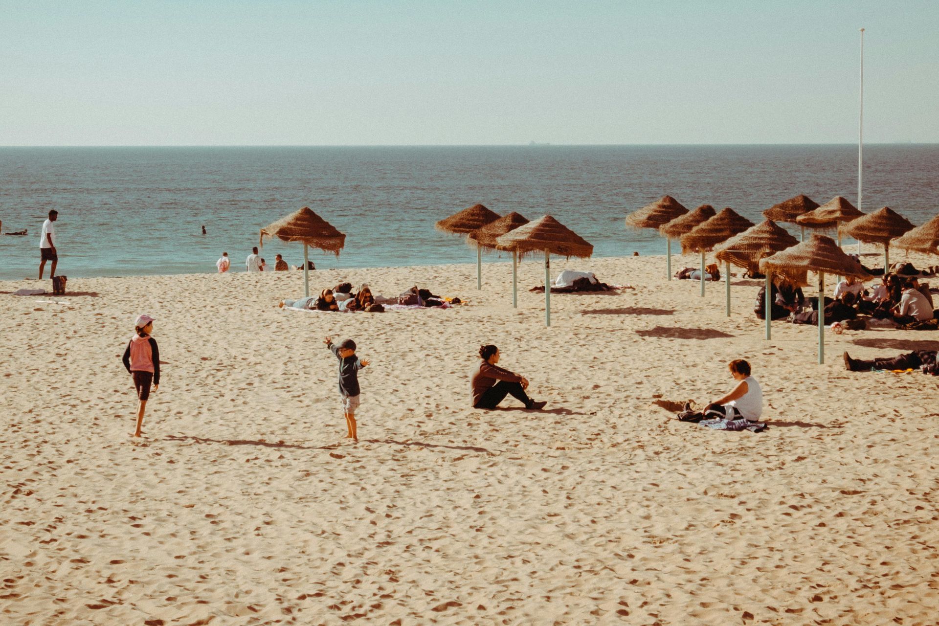 Praia de areia com guarda-sóis de palha, pessoas tomando sol e nadando. Oceano azul sob um céu limpo.