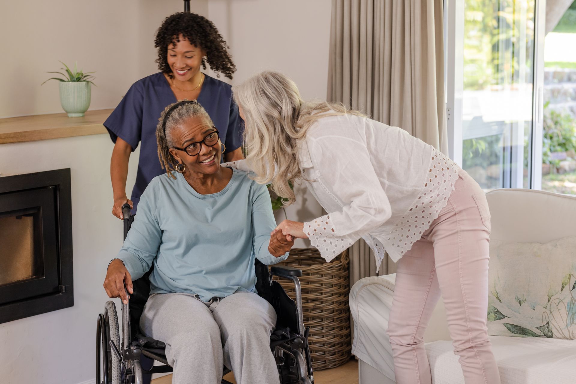 Une femme en fauteuil roulant sourit, une autre femme l'aide à se lever, une soignante est visible en arrière-plan