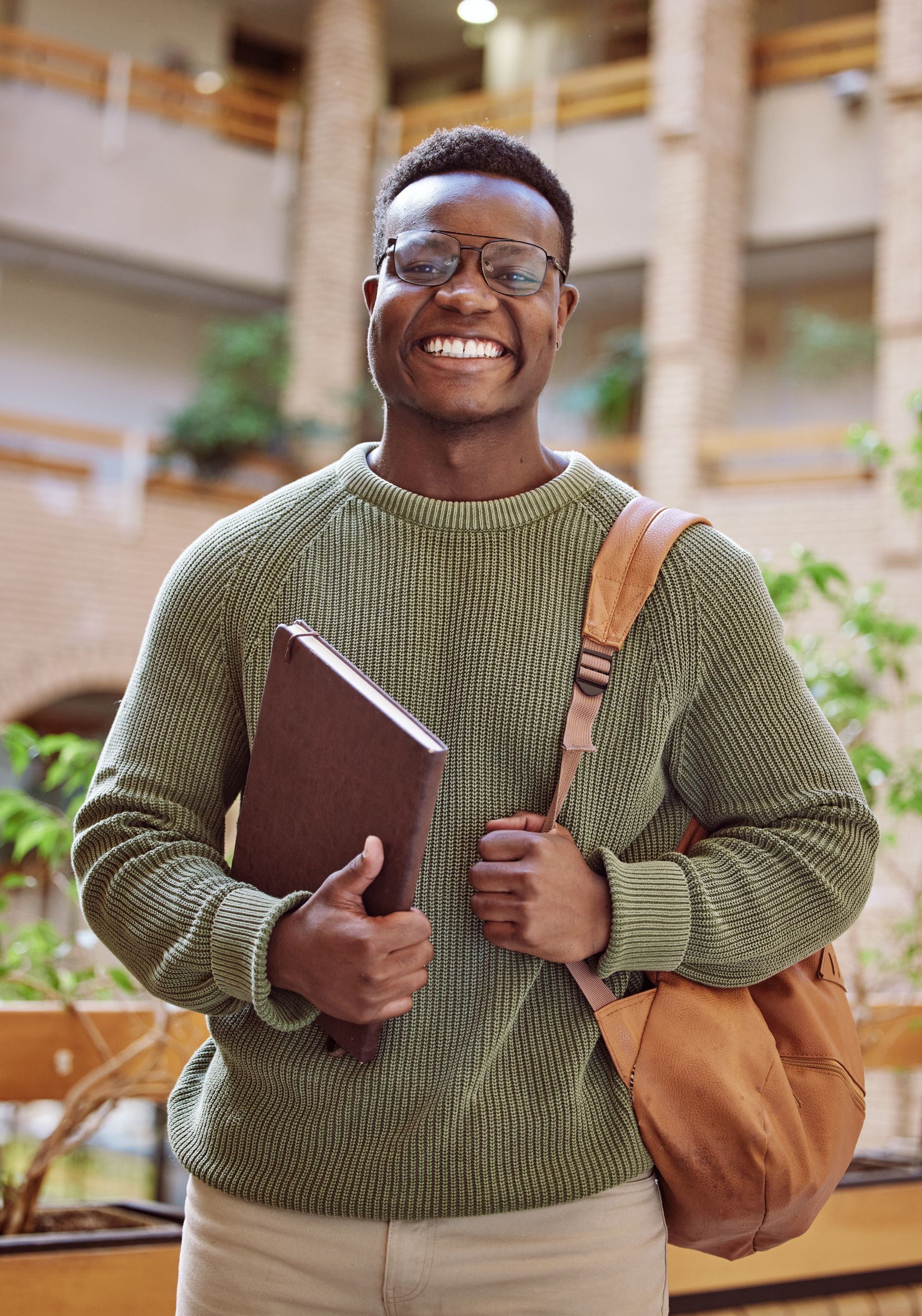 Personne souriante portant des lunettes, tenant un livre et un sac bandoulière, debout dans un bâtiment entouré de verdure