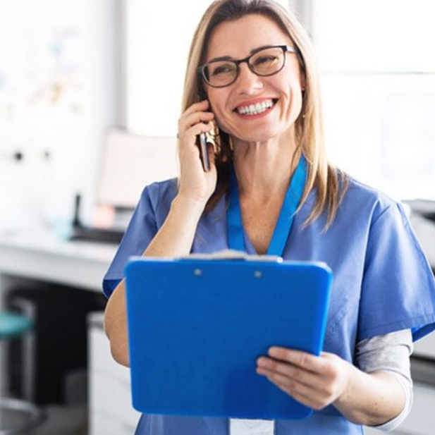 Une infirmière souriante en tenue bleue parle au téléphone tout en tenant un presse-papiers
