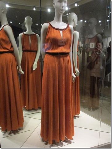Des mannequins en robes orange à bordures noires, exposés sous verre.