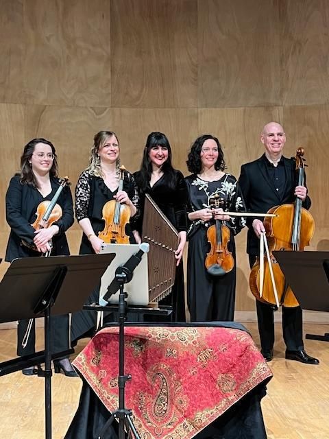 Un ensemble musical de cinq personnes tenant des instruments, debout sur scène, souriantes.