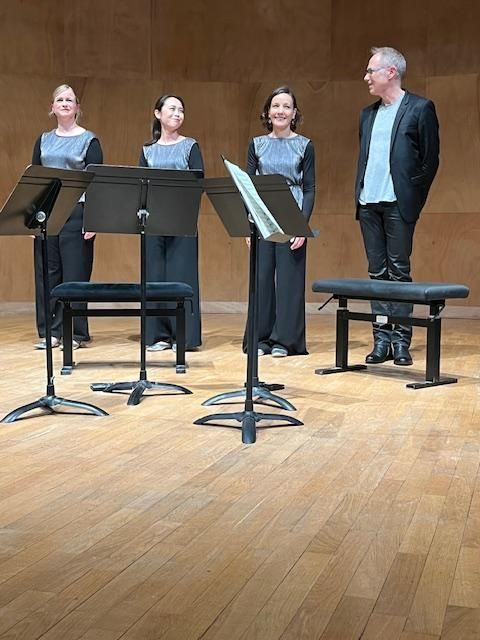 Un chœur sur scène, trois femmes et un homme, avec des partitions.