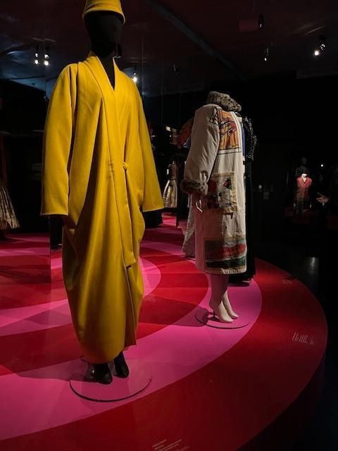 Robe-manteau jaune et manteau à motifs sur des mannequins dans une exposition muséale aux sols roses et rouges.