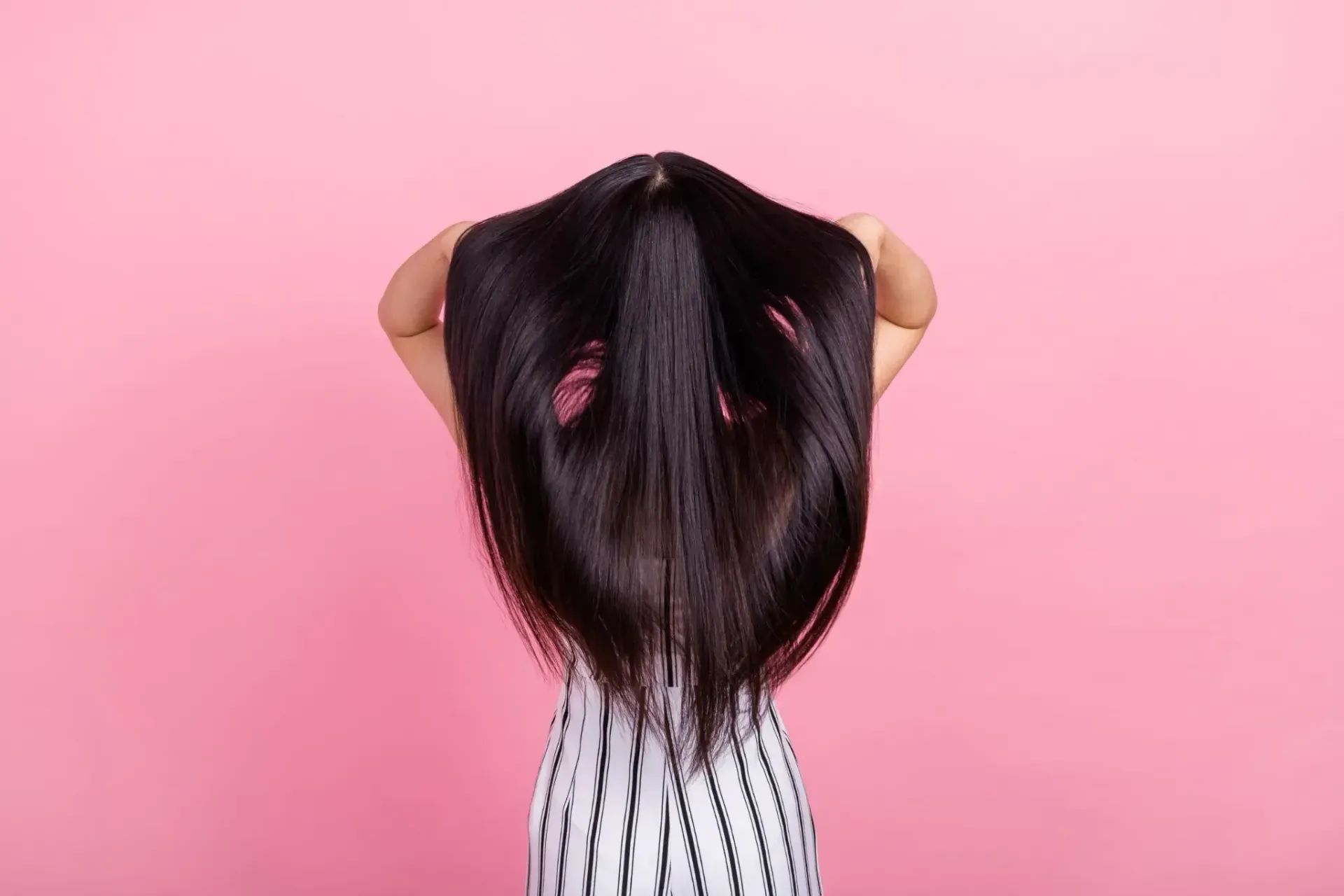 Mujer con cabello largo y negro, con los brazos en alto, de pie sobre un fondo rosa.