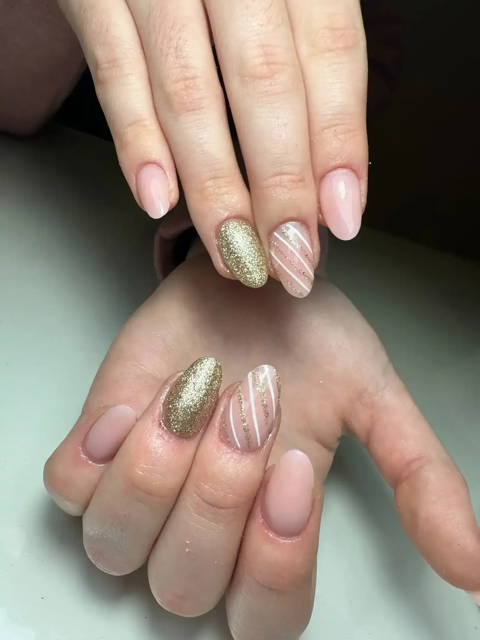 Primer plano de manos con uñas ovaladas, decoradas con purpurina rosa claro y dorada. Dos uñas tienen rayas blancas.