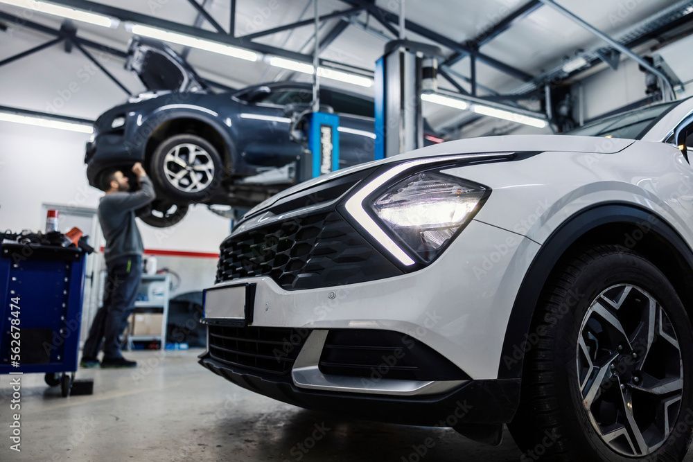 Ein Automechaniker arbeitet in einer Autowerkstatt unter einem hochgehobenen blauen SUV neben einem weissen SUV.