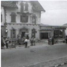 Schwarz-Weiss-Strassenszene mit einem zweistöckigen Gebäude, davor stehenden Personen und einem in der Nähe geparkten Kleinwagen.