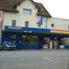 Peugeot-Autohaus mit blauer Ladenfront an einer Strasse, einem davor geparkten blauen Auto und einem darüberliegenden Gebäude.