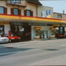 Strassenansicht eines kleinen Ladengeschäfts mit gelb-roter Markise und davor geparkten Autos.
