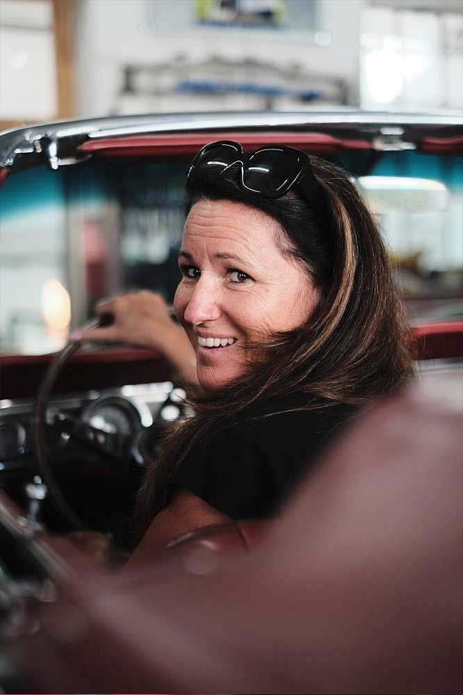 Frau lächelt in einem roten Oldtimer, Sonnenbrille auf dem Kopf, Lenkrad sichtbar.