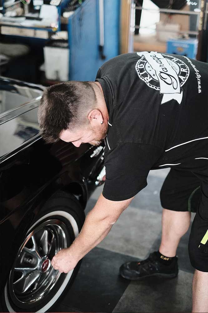 Ein Mechaniker beugt sich in einer Garage über das Rad eines schwarzen Autos, um es zu inspizieren.