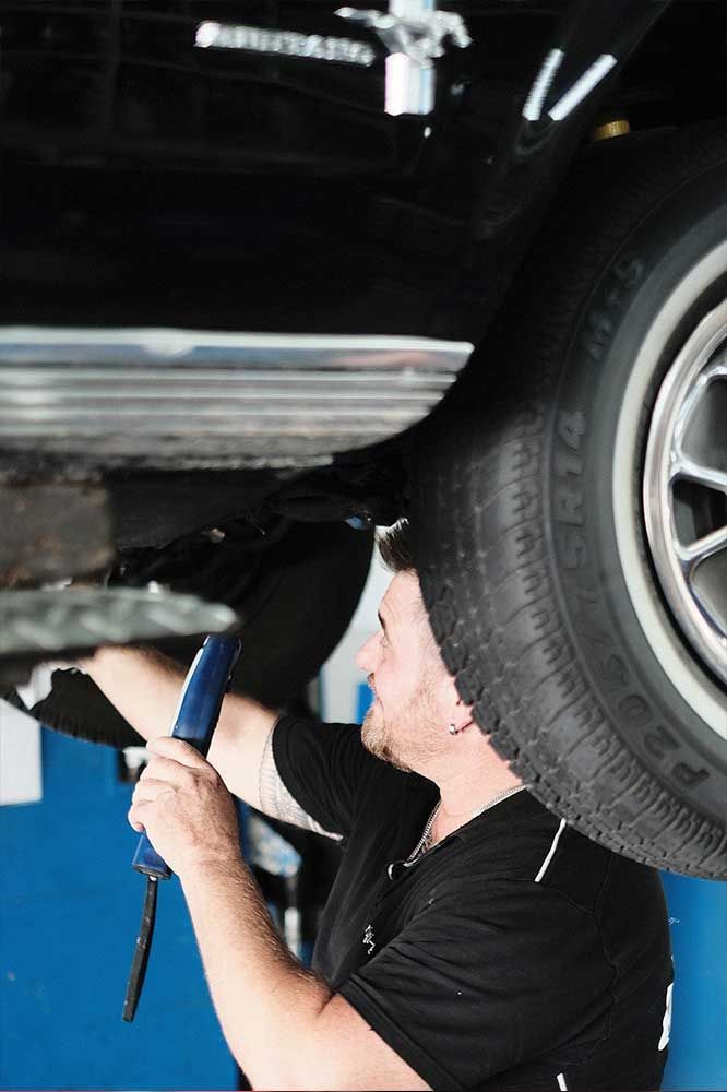 Ein Mechaniker untersucht in einer Garage ein auf einer Hebebühne stehendes Auto mit einer Taschenlampe.