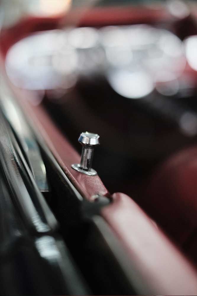 Nahaufnahme einer kleinen silbernen Schraube auf einer roten Metalloberfläche; im Hintergrund ist der Innenraum eines Autos verschwommen zu sehen.